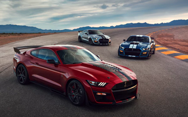 Silver, red, and blue Ford Mustang Shelby GT500 muscle cars are positioned on a desert racetrack under a cloudy sky in this 4K Ultra HD desktop wallpaper.