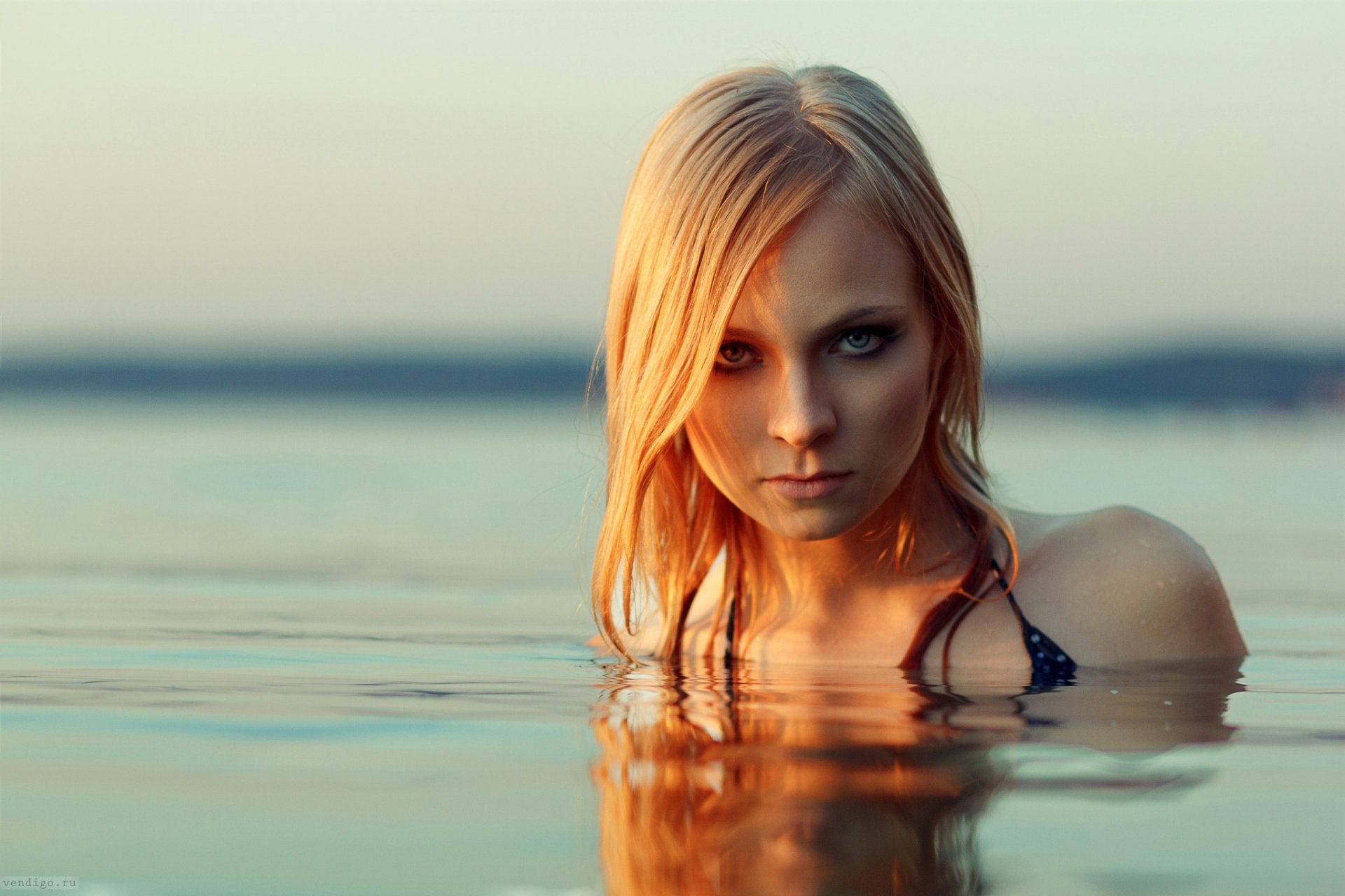 Blonde woman with blue eyes stares intensely while partially submerged in calm water, creating a reflective effect in this HD desktop wallpaper.