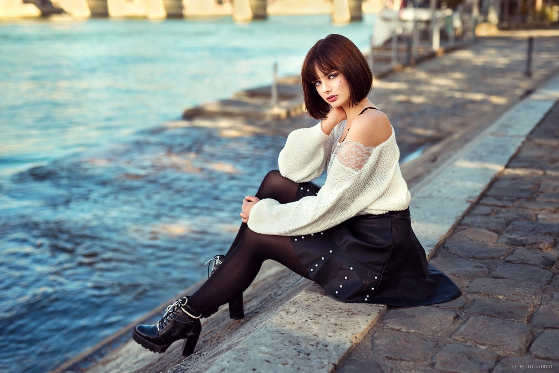 French model Marie Grippon with short brunette hair sits by the water wearing a skirt, black tights, and a white off-shoulder sweater in this HD desktop wallpaper.