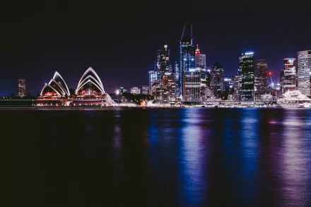 Nighttime view of Sydney city skyline featuring the illuminated Sydney Opera House and high-rise buildings, reflected in calm waters. 4K Ultra HD desktop wallpaper.