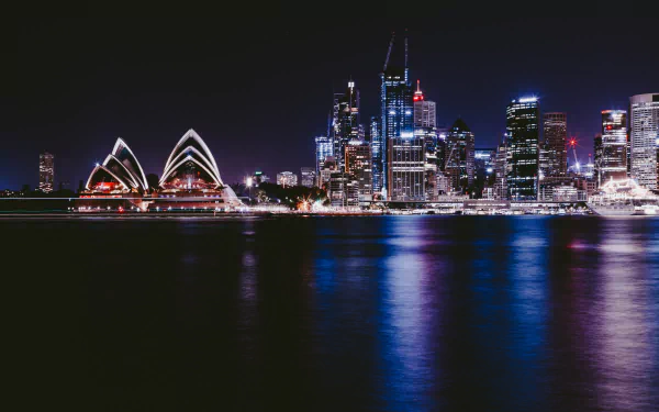 Nighttime view of Sydney city skyline featuring the illuminated Sydney Opera House and high-rise buildings, reflected in calm waters. 4K Ultra HD desktop wallpaper.