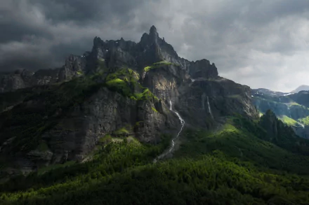 HD desktop wallpaper of a dramatic mountain peak with a cascading waterfall flowing down a steep cliff surrounded by lush green nature under moody clouds.